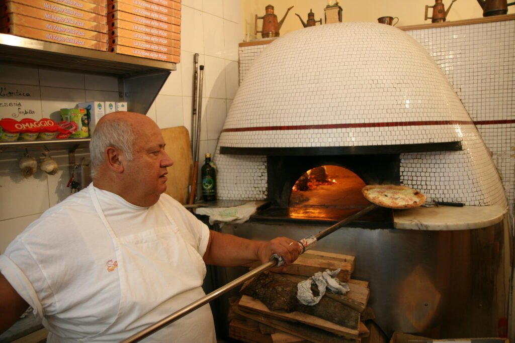 Pizzaiolo com uma pizza na pá diante de um forno a lenha em Nápoles